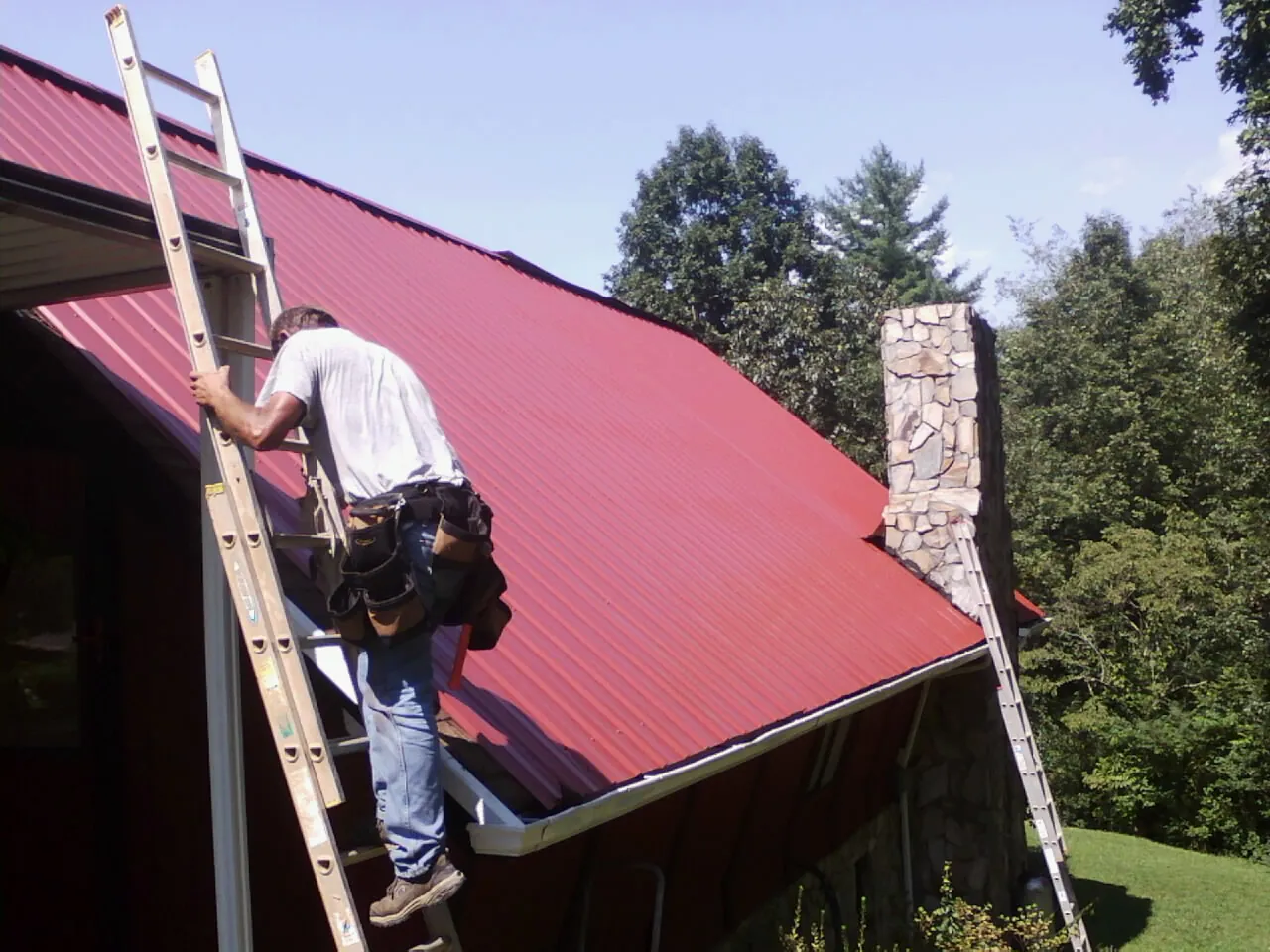 Roofer on a ladder beside a red metal roof and stone chimney — Soffit & Fascia Repair by Roofing Brothers in Contoocook